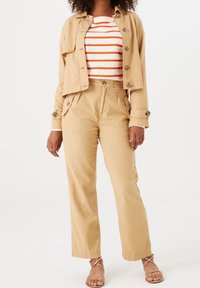 Beżowa krótka kurtka z guzikami, zestawiona z beżowymi spodniami z wysokim stanem i w paski bluzką w czerwono-białe pasy. Całość dopełniają sandały.