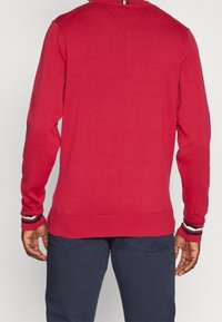 Pull rouge en tricot avec manches longues, poignets rayés contrastants et col en V. Texture lisse et coupe ajustée.