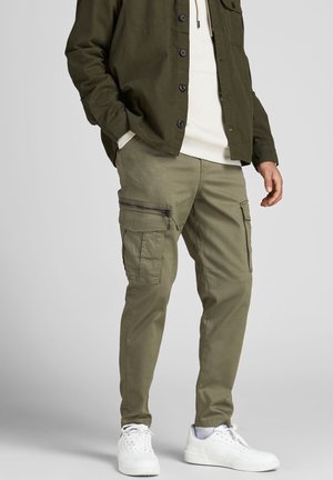 Olivenfarbene Cargo-Hosen mit mehreren Taschen und Reißverschlus Akzenten, kombiniert mit einem leichten Hoodie und einem dunklen Hemd mit Knöpfen. Weiße Sneakers runden den Look ab.