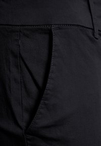 Pantaloni neri in tessuto presentano una superficie texturizzata con dettagli di cucitura e una tasca laterale. Il design è elegante e sagomato per un look aderente.