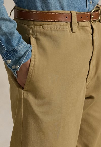 Main dans la poche d'un pantalon beige avec une ceinture en cuir marron, associée à une manche de chemise en denim bleu clair avec boutons-pression métalliques.
