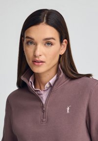 Sudadera de forro polar morada con cremallera y un alto cuello, que presenta un logo de color claro y una camisa de cuadros rosa y blanca a rayas debajo.