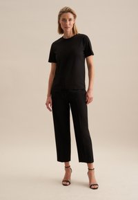 Zwart T-shirt met korte mouwen, gecombineerd met zwarte cropped broek en zwarte sandalen met bandjes, weergegeven tegen een neutrale achtergrond.