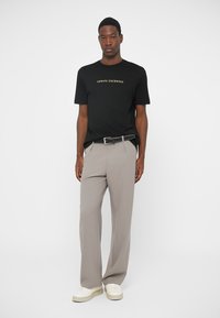 Άντρας φοράει μαύρο μπλουζάκι Armani Exchange, γκρι παντελόνι με μαύρη ζώνη και λευκά αθλητικά παπούτσια, μπροστά σε μονόχρωμο ανοιχτό φόντο.