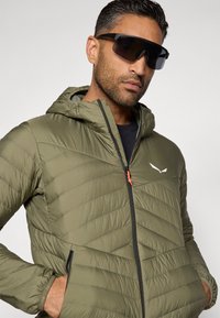 Casaco acolchoado verde-oliva com padrão em chevron, frente com fecho éclair e capuz. Apresenta um puxador de fecho e um logotipo em laranja contrastante.