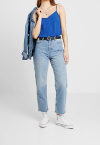 Žena v modré košili s puntíky, světle modrých vysokých džínách, bílých teniskách s vysokým svrškem, držící denimovou bundu přes jedno rameno.