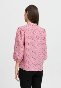 Rosa Bluse aus strukturiertem Stoff, mit einem Muster aus kleinen schwarzen Herzen. Puffärmel und ein runder Ausschnitt runden das Design ab.