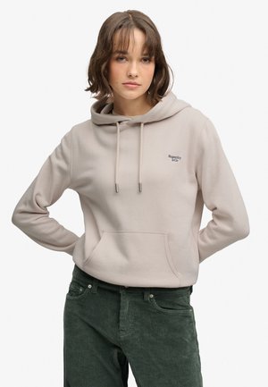 Giovane donna con capelli castani che indossa una felpa beige con cappuccio e pantaloni verdi in velluto a coste, in piedi con le mani dietro la schiena.