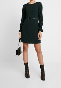 Svart långärmad klänning med rundad halsringning, åtsittande midja med ett tunt bälte, matchad med texturerade svarta ankelstövlar och en liten svart handväska.