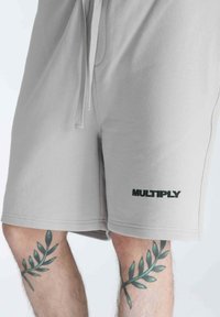 Shorts in cotone grigio chiaro con vita regolabile, caratterizzati da un logo nero "MULTIPLY" sulla gamba inferiore e una texture liscia e morbida.