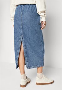 Jupe midi en denim bleu moyen, avec une fente haute à l'arrière, une taille élastique et un ourlet effiloché. Associée à des baskets blanches et des chaussettes.