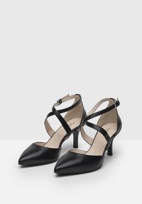 Talons en cuir noir avec un bout pointu, comportant des brides entrecroisées et un talon de 5 cm. Texture lisse et design minimaliste.
