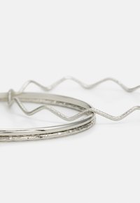 Două accesorii pentru păr metalice de argint: unul neted și rotund, celălalt ondulat cu o finisare texturată. Sunt împletite pe un fundal alb.