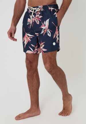 Mandlig model iført marineblå badeshorts med pink og hvidt tropisk bladtryk, elastisk linning, hvid snor, står barfodet.