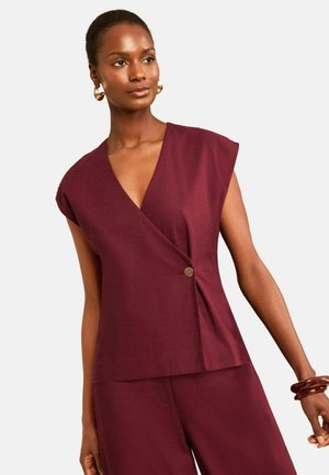 Vrouw draagt een mouwloze bordeaux wikkeltop met één knoop en bijpassende wijde broek, geaccentueerd met gouden creolen en armbanden.