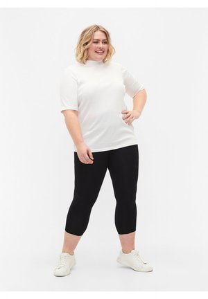 Smaidoša sieviete stāv, roka uz gūžas, tērpusies baltā īsroku krekliņā, melnās sasprostotās legingās un baltos sporta apavos uz vienkārša fona.