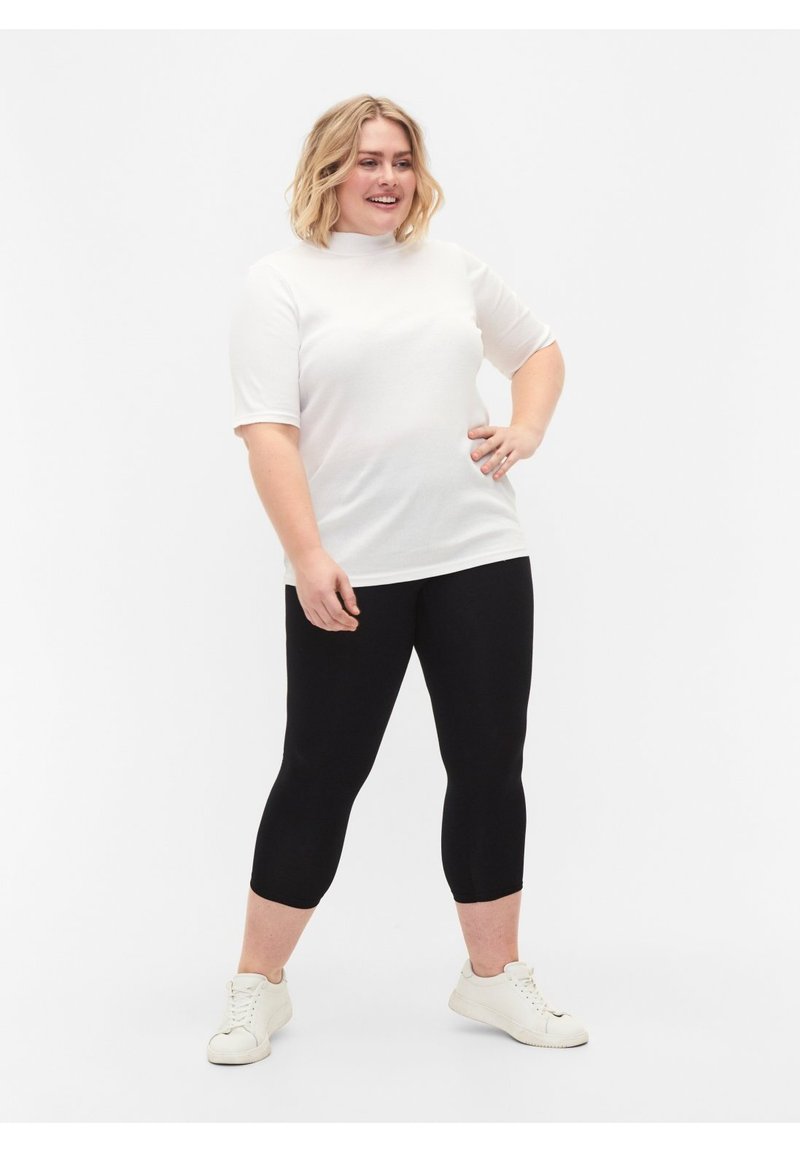 Lachende vrouw staat met hand op heup, draagt een wit T-shirt met korte mouwen, zwarte cropped leggings en witte sneakers tegen een effen achtergrond.