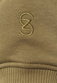 Primer plano de un suéter caqui que presenta un tejido de punto suave, un dobladillo acanalado y un logo bordado en oro en la parte frontal.