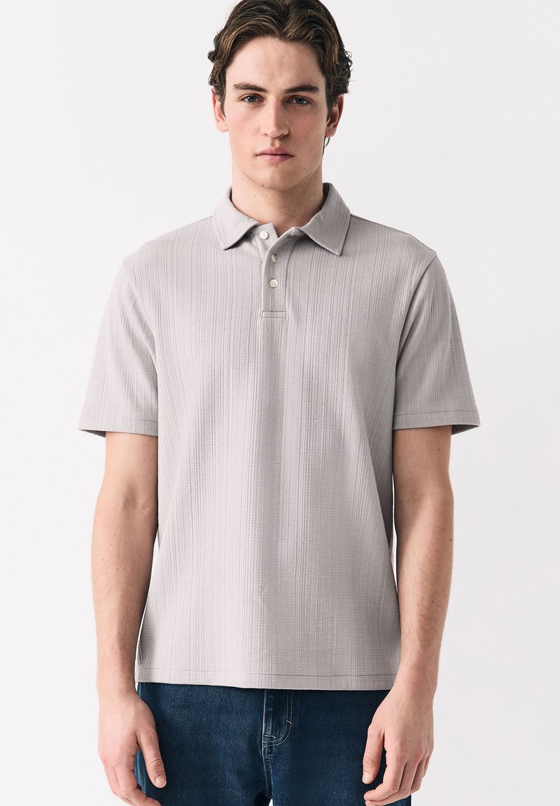 Giovane uomo che indossa una polo a maniche corte grigio chiaro con trama e jeans blu scuro, in piedi davanti a uno sfondo bianco semplice.