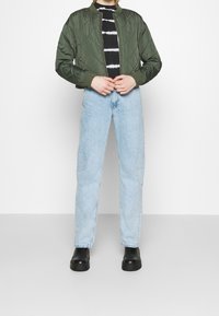 Zielona pikowana kurtka bomberka z prążkowanymi mankietami, noszona na czarno-białym paskowanym topie, w połączeniu z jasnoniebieskimi jeansami o prostych nogawkach i czarnymi botkami.