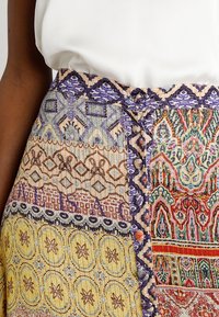 Veelkleurige rok met patroontjes in blauw, geel en rood, met een textuurstof en decoratieve knopen.