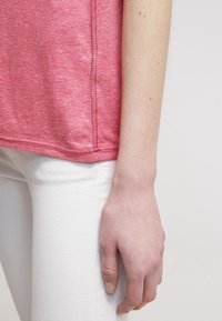 Rechterarm van een persoon hangt naast het lichaam, gekleed in een roze shirt en witte broek, tegen een eenvoudige lichte achtergrond.