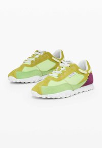 Zelené a žluté semišové tenisky s bílou podrážkou, s fialovými akcenty, vrstveným designem a bílými tkaničkami. Texturované povrchy a detaily šití.