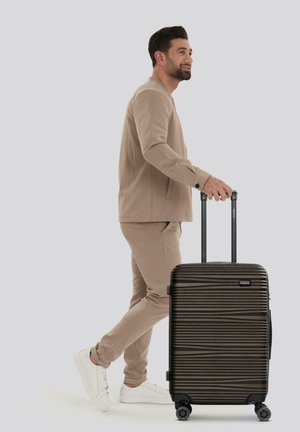 Valigia nera compatta con scanalature orizzontali, doppie ruote e manico telescopico. Il modello indossa un outfit beige e sneakers bianche.