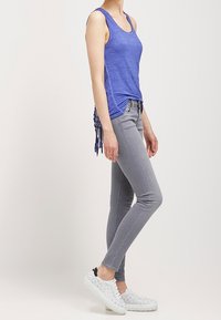 Top sin mangas azul con drapeado lateral, combinado con jeans ajustados gris claro y zapatillas blancas con patrón.