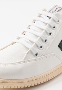 Białe sneakersy z teksturowanym syntetycznym wierzchem, okrągłym noskiem, białymi sznurówkami, zielonymi akcentami oraz beżową gumową podeszwą o teksturowanym wykończeniu.