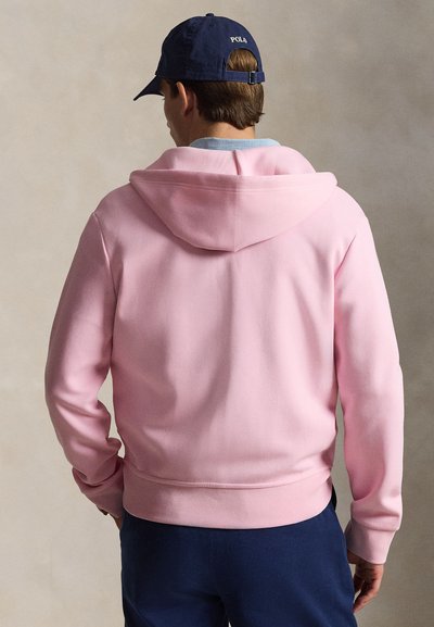 Mees, kes on seljaga, kannab heleroosat kapuutsiga dressipluusi, tumesiniseid pükse ja tumesinist nokamütsi, millel on taga tikandiga "POLA".