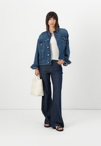 Denimjakke med to framlommer, paired med mørkeblå utsvingte jeans. Har en teksturert hvit veske, iført svarte sandaler. Enkel, casual stil.