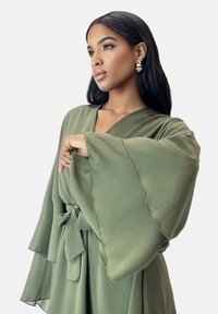 Olivgrünes Kleid im Kimono-Stil mit weiten, mehrlagigen Ärmeln und tailliertem Gürtel. Glatter, leichter Stoff. Ohne Muster oder Verzierungen.