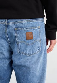 Tagantvaade inimesest, kellel on seljas helesinised teksad, millel on nahast Carhartti logo silti tagakotil, ja must pikkade varrukatega särk.