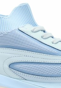 Zapatilla deportiva azul claro con tejido de punto, superposiciones de cuero y cordones azul claro, vista en primer plano enfocándose en el lateral y la parte superior.