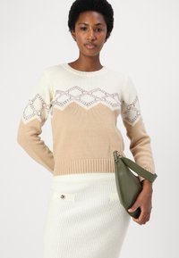 Maglione lavorato a maglia beige e crema con motivo geometrico in pizzo, polsini e orlo a coste. Abbinato a una borsa a mano in pelle verde.