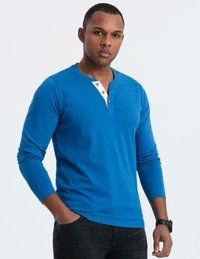 Man som bär en blå långärmad henley-tröja med vit knappslå, svarta jeans och en svart armbandsklocka, står med en hand i fickan.