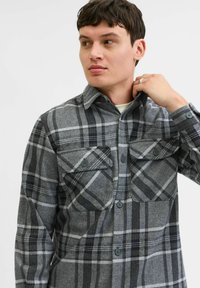 Camisa de franela a cuadros grises con grandes bolsillos en el pecho, cierre de botones en el frente y cuello. La tela tiene una apariencia suave y texturizada.