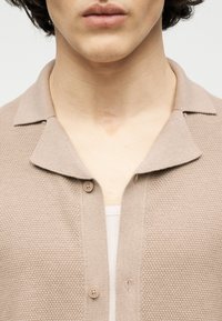 Κοντινό πλάνο ενός ατόμου που φοράει μια μπεζ πλεκτή ζακέτα με υφή πάνω από μια λευκή ριγωτή μπλούζα, εστιάζοντας στον λαιμό και την περιοχή του γιακά.