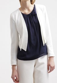 Weißer kurzer Blazer aus strukturiertem Stoff, kombiniert mit einer marineblauen Bluse mit Faltendetails, getragen über weißen Hosen.