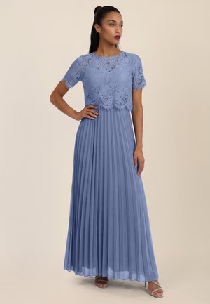 Frau trägt ein bodenlanges, staubblaues Kleid mit kurzen Spitzärmeln und einem plissierten Rock, steht mit einer Hand in der Hüfte und schaut seitlich.