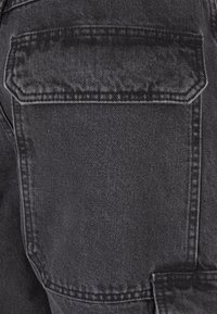 Crni denim materijal sa glatkom teksturom sadrži stražnji džep, ističući detaljno šivanje i čist, minimalistički dizajn.