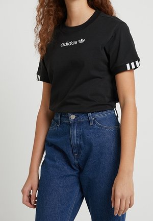 Frau trägt ein schwarzes Adidas-T-Shirt mit weißem Logo und Streifen an den Ärmeln, kombiniert mit hoch taillierter blauer Jeans, steht vor einfarbigem Hintergrund.