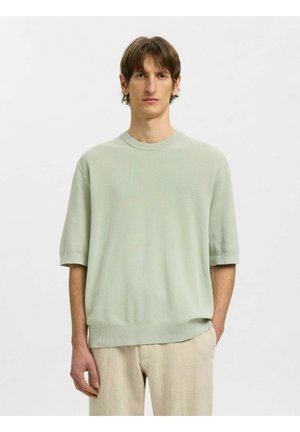 Mann trägt hellgrünen kurzärmeligen Strickpullover und beige Hose, steht vor einfachem weißen Hintergrund.