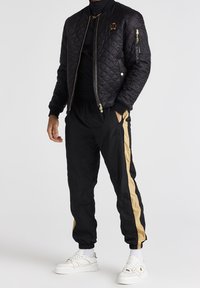 Svart quiltad bomberjacka med guldaccenter och en badge, tillsammans med svarta byxor med en guldstrimma, och vita sneakers.