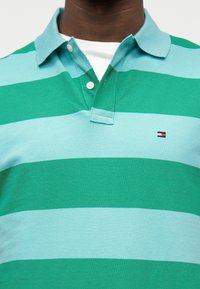 Άνδρας που φοράει πράσινη και γαλάζια ριγέ πόλο μπλούζα με δύο κουμπιά, λευκό μπλουζάκι να φαίνεται στον γιακά και μικρό λογότυπο στο στήθος.