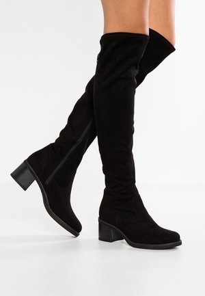 Bottes noires en daim montantes jusqu'aux genoux, avec talons carrés et fermetures éclair latérales, portées sur des jambes croisées sur un fond blanc.