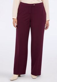 Woman wearing high-waist, wide-leg burgundy trousers with a buttoned waistband and beige heeled shoes against a plain background.