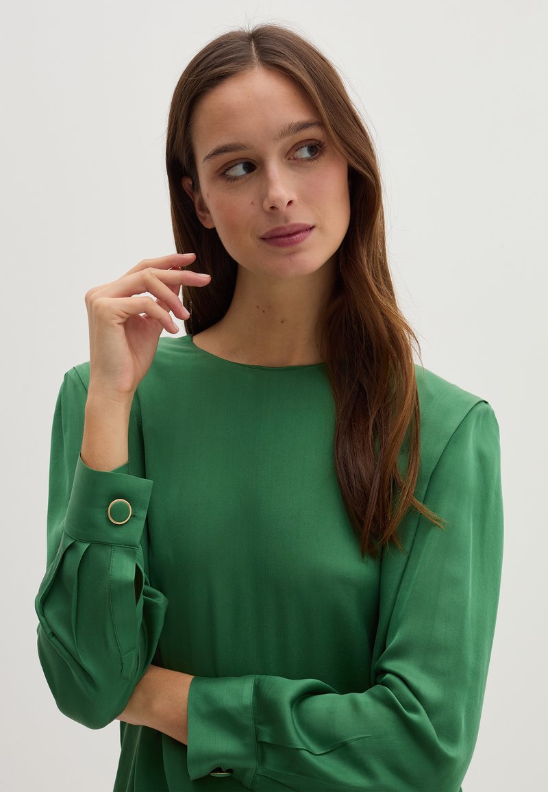 Grüne Bluse mit glatter, satinhafter Textur. Verfügt über lange Ärmel mit Knopfdetails und einen runden Halsausschnitt. Einfaches, elegantes Design.