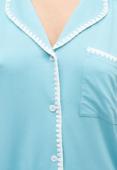 Chemise bleu clair à boutons avec bordure blanche en forme de coquilles sur le col, la ligne de boutons et la poche poitrine.